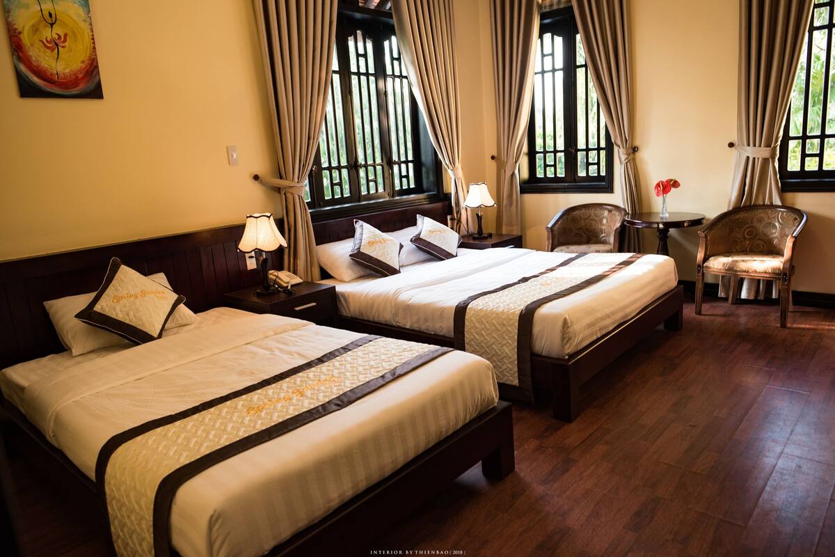 Quarto de hotel com duas camas, uma mesinha com abajur, uma cadeira e um quadro na parede.