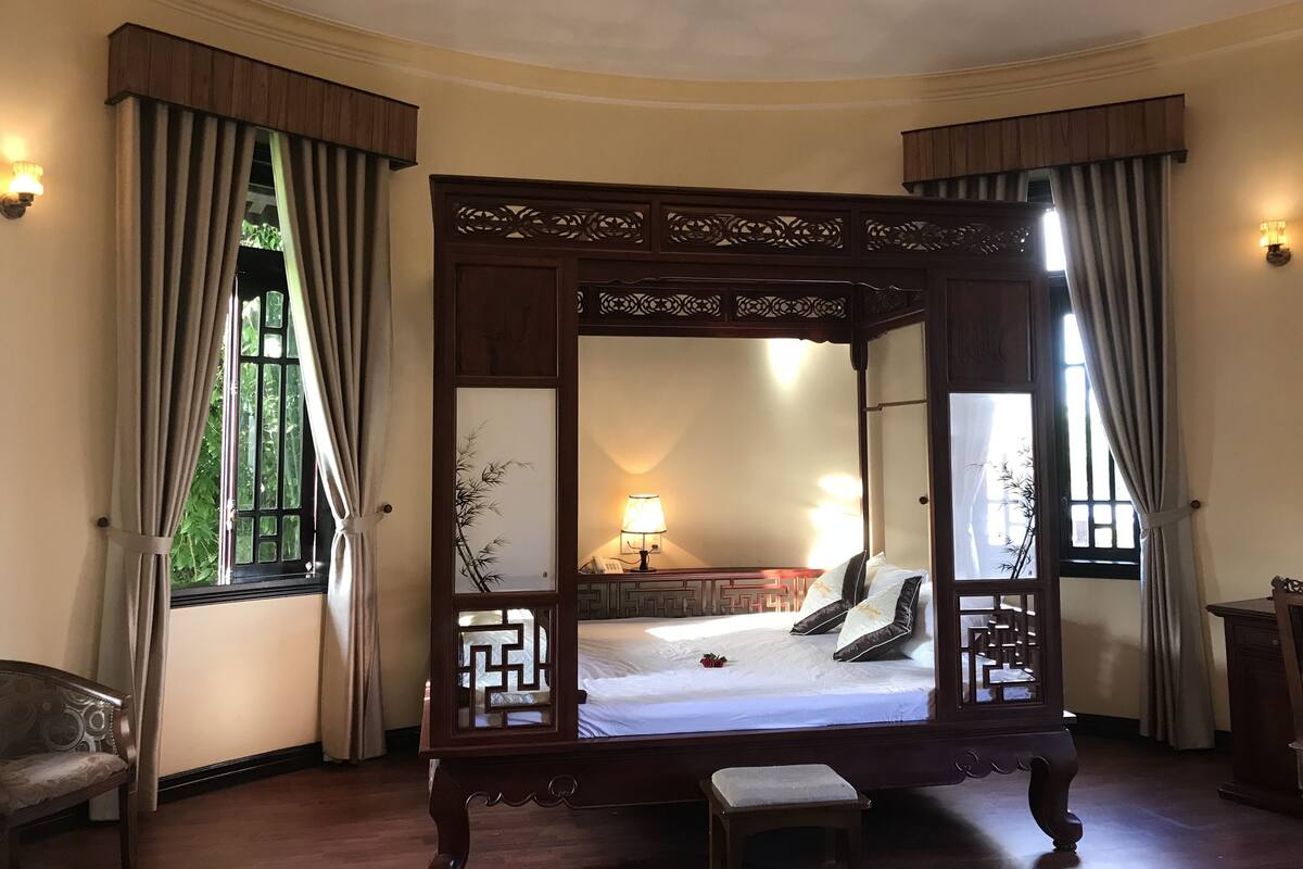 Quarto tradicional com cama de dossel, mesa de cabeceira pequena e uma cadeira.