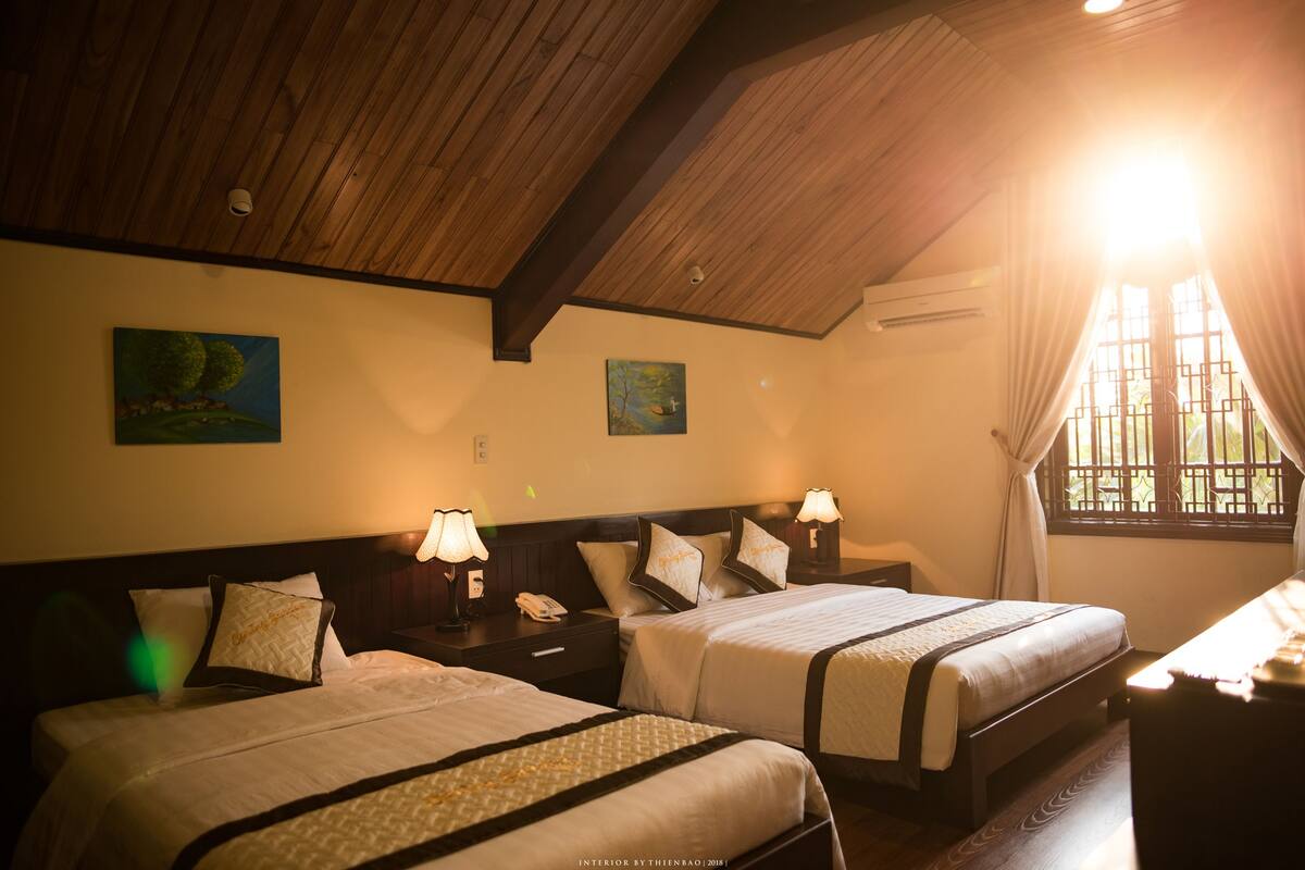 Quarto de hotel com duas camas, vigas de madeira no teto, obras de arte nas paredes e uma janela com cortinas.