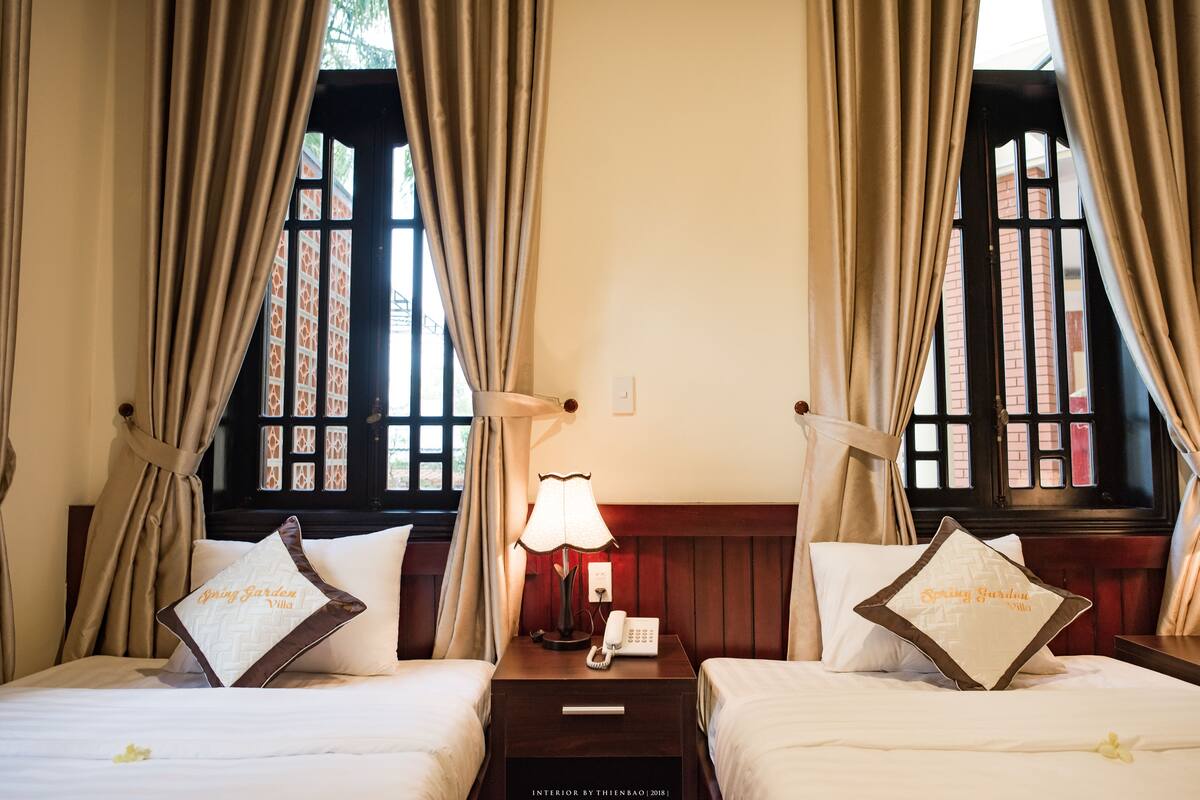 Quarto de hotel com duas camas, uma mesa de cabeceira com abajur e uma janela com cortinas.