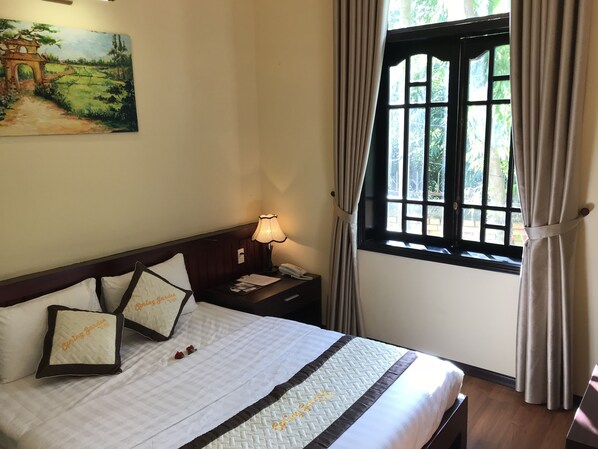 A hotel room with a bed, a nightstand, a lamp, a window with curtains, and a painting on the wall.