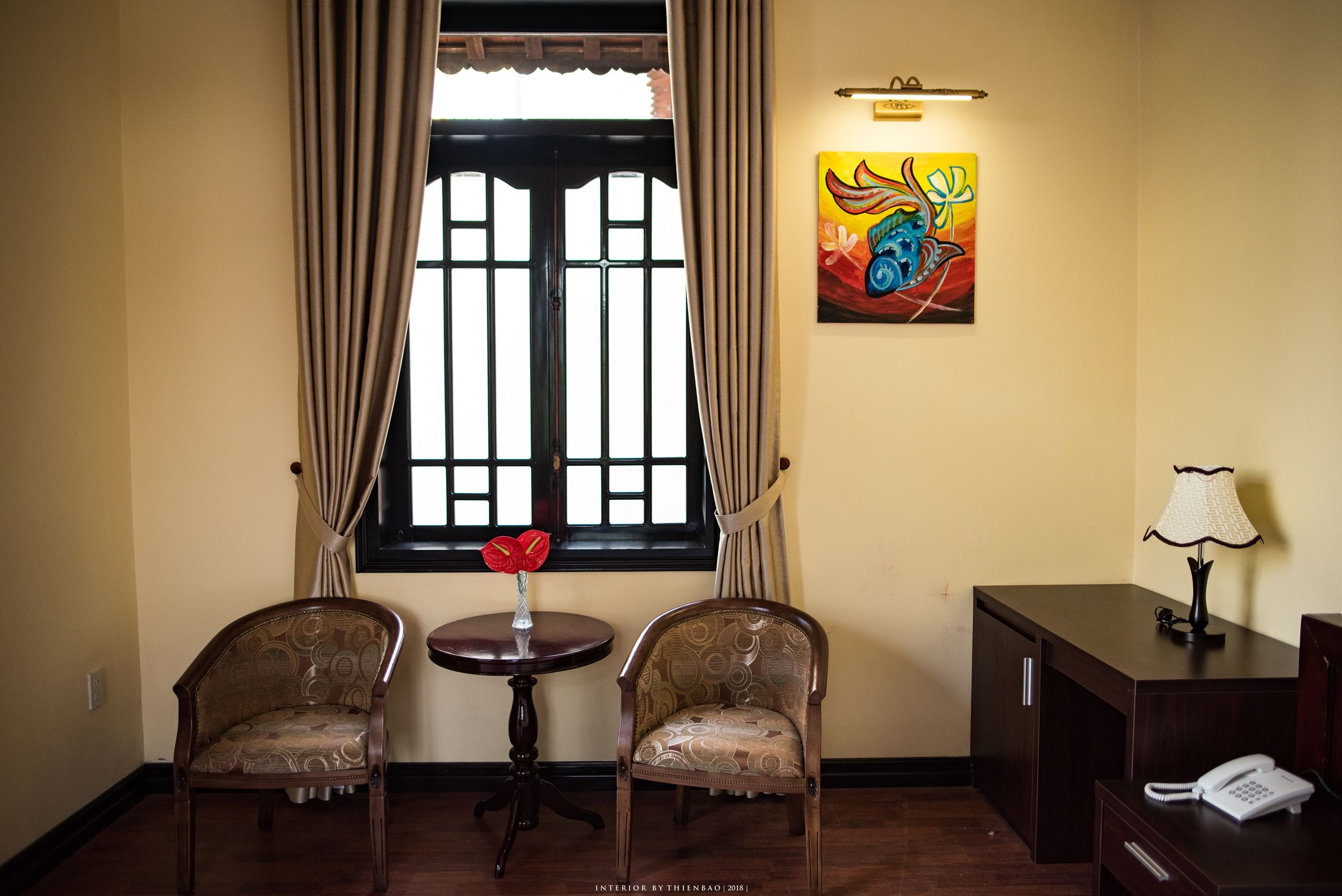 A room with two chairs, a small table with a vase, a painting, a lamp, and a phone on a desk.