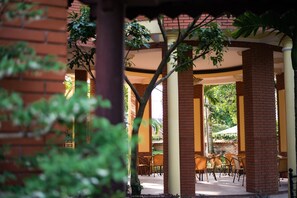 A covered outdoor seating area with brick pillars and wooden chairs, surrounded by greenery.