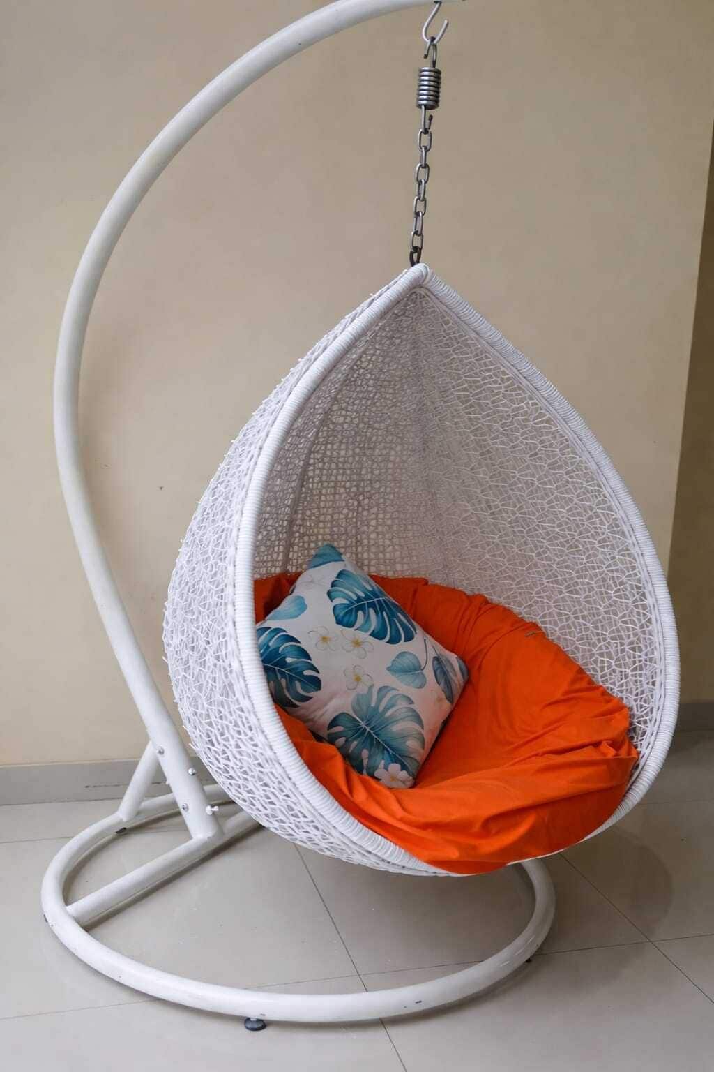 En vit hängstol med ett säte av nätmaterial och en orange kudde, samt en prydnadskudde med blommotiv.