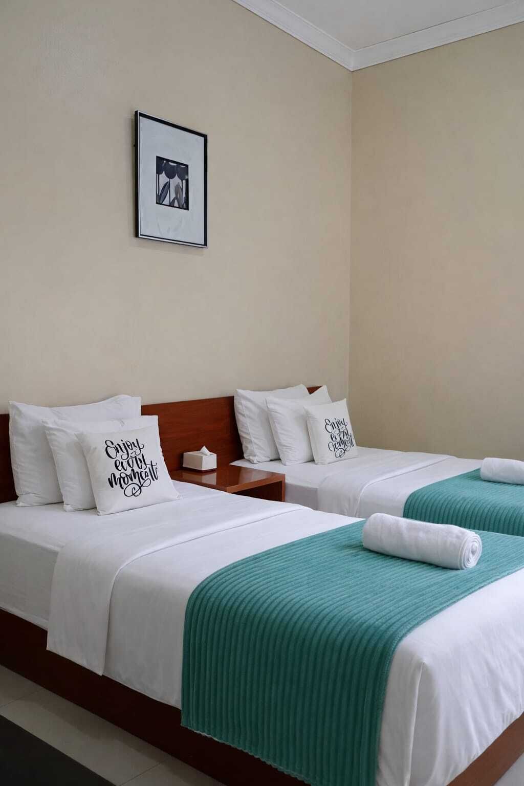 A hotel room with two beds, each with white bedding and teal throw blankets. There are white pillows with text, a framed picture on the wall, and a wooden headboard.