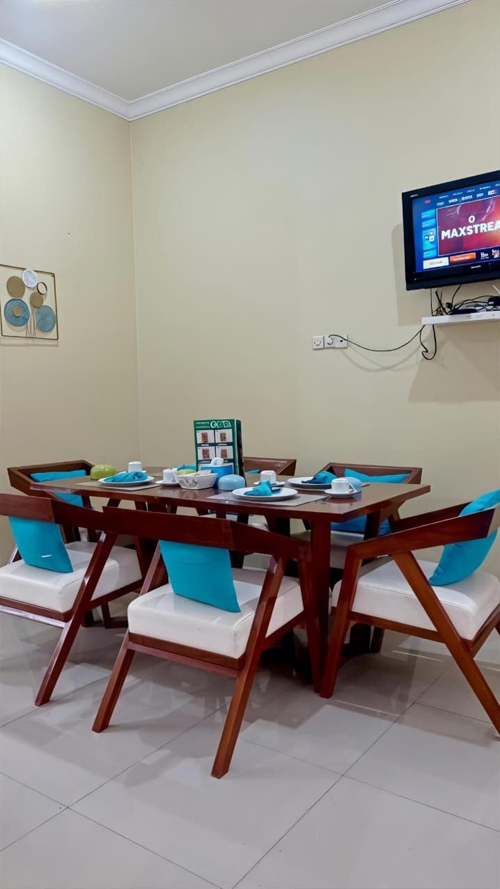 A dining area with a round table and six chairs, a television mounted on the wall, and a painting on the wall.
