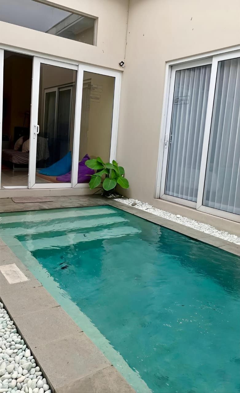 Una piscina con borde de baldosas, rodeada por un muro bajo y un pequeño jardín con guijarros blancos y plantas verdes.