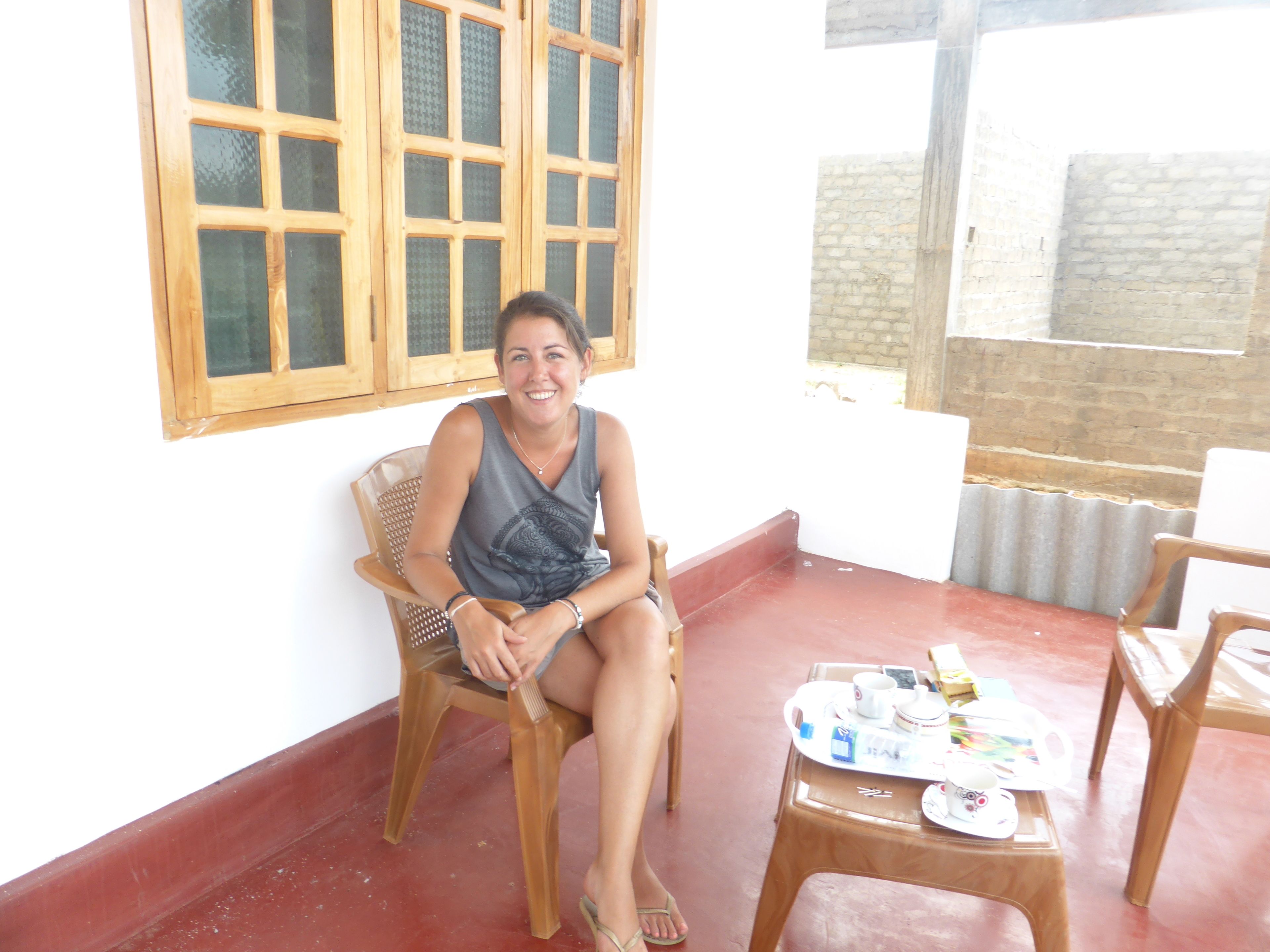 Une personne assise sur une chaise en bois, dans une pièce avec une fenêtre, une table sur laquelle se trouve un plateau rempli d’objets, et un mur en briques.