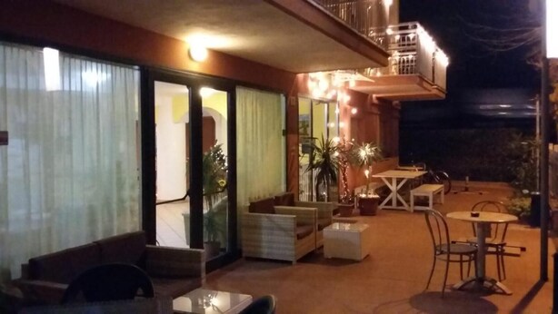 An outdoor patio area with seating and tables, a balcony with lights, and a view of the street. - Hotel Frida (Rimini)