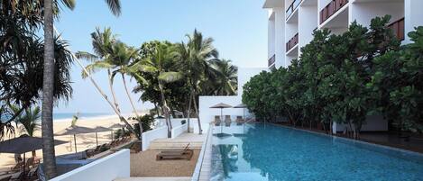 Área de piscina en un resort, con vista despejada al océano, rodeada de palmeras y un edificio moderno con balcones.
