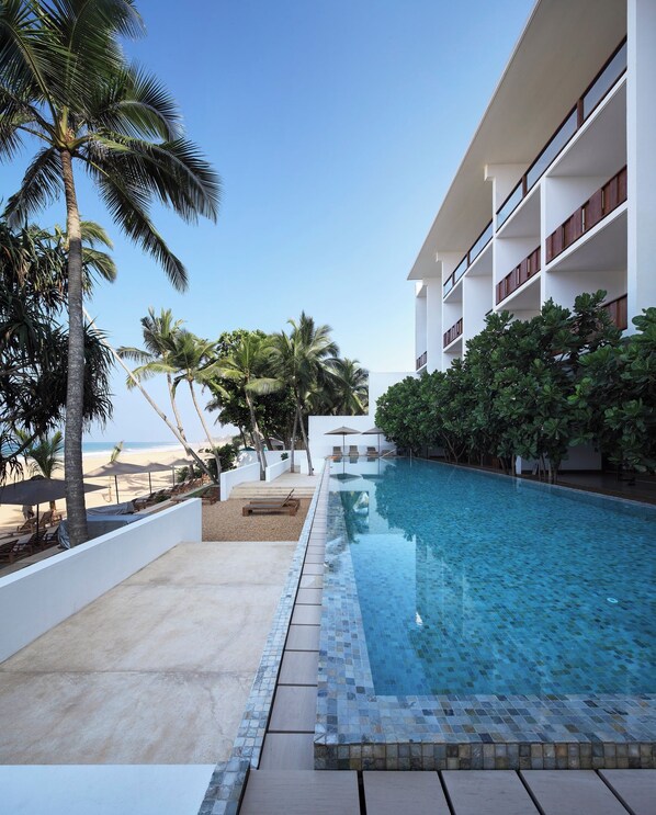 A resort pool area with a clear view of the ocean, surrounded by palm trees and a modern building with balconies.