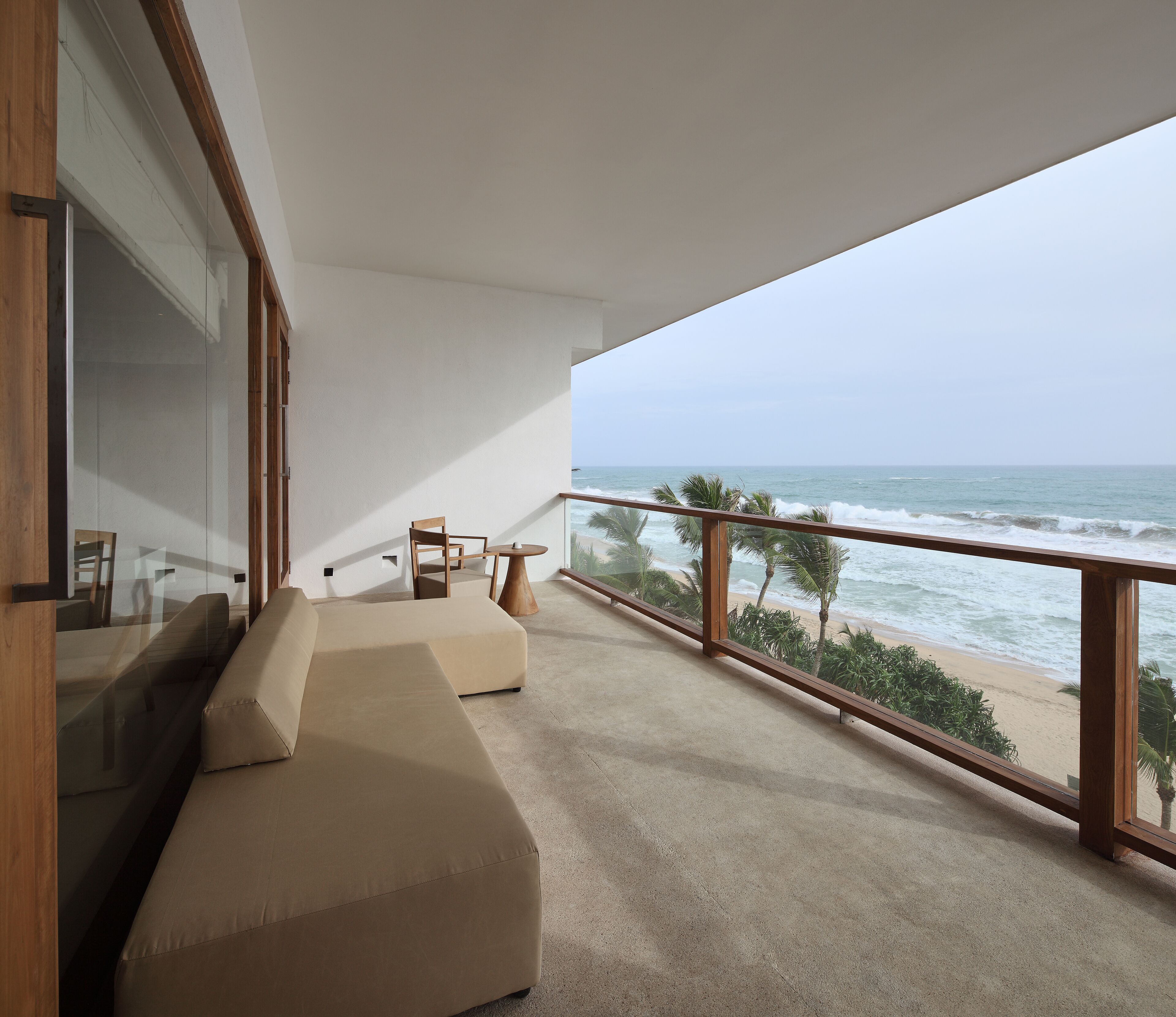 A balcony with a beige sofa and chairs, overlooking a beach with palm trees and the ocean.