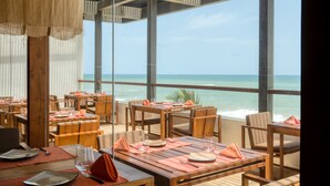 Een restaurant aan zee met houten tafels en stoelen, grote ramen en uitzicht op de oceaan.