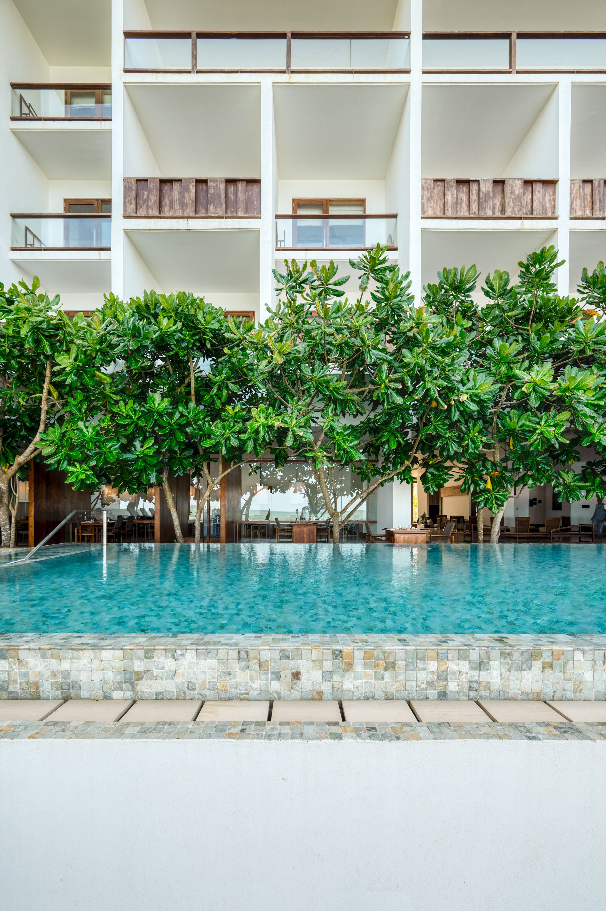 A pool area with a tiled edge, surrounded by lush green trees and a multi-story building with balconies.