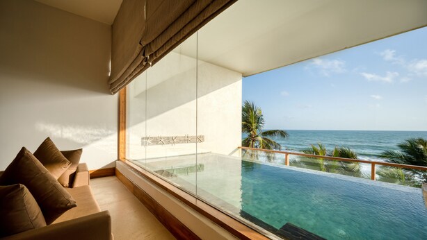 Een balkon met glazen rand, met uitzicht op een heldere blauwe oceaan en palmbomen.
