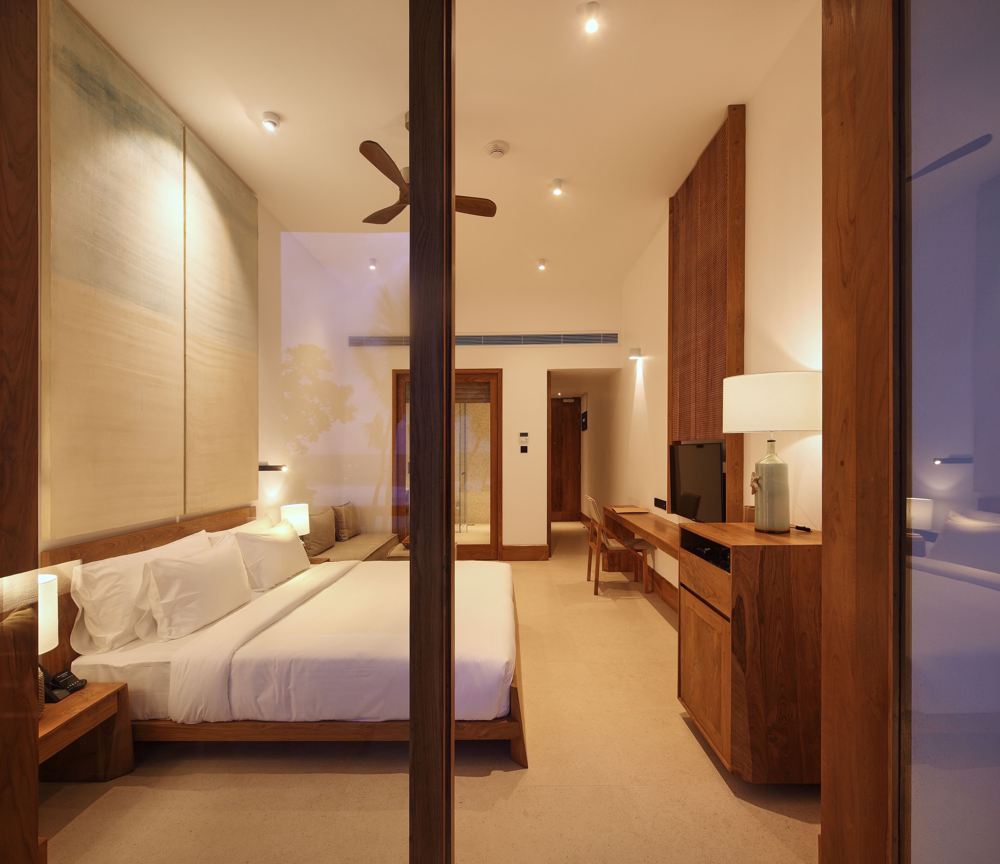 A modern hotel room with a large bed, a desk, a television, and a ceiling fan.