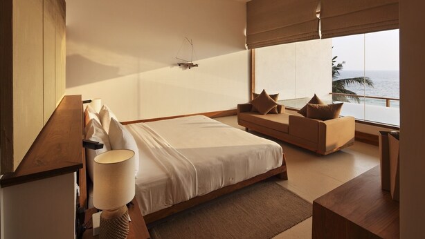A modern bedroom with a large bed, a sofa, a ceiling fan, and a view of the ocean.