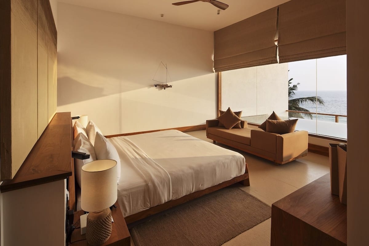 Quarto moderno com uma cama grande, um sofá, um ventilador de teto e vista para o mar.