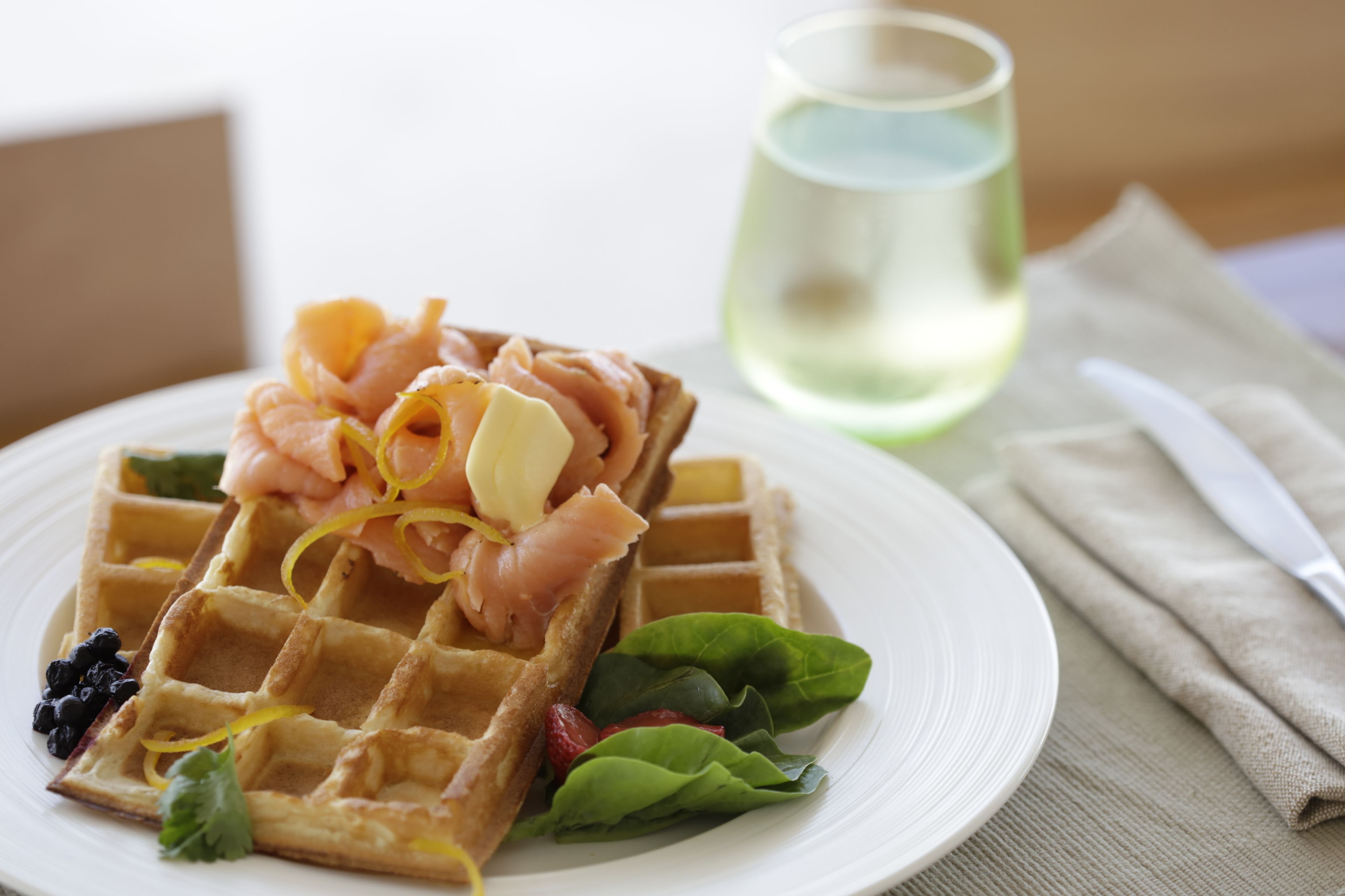 A plate with a waffle topped with salmon, cheese, and greens, a glass of water, and a knife on a napkin.