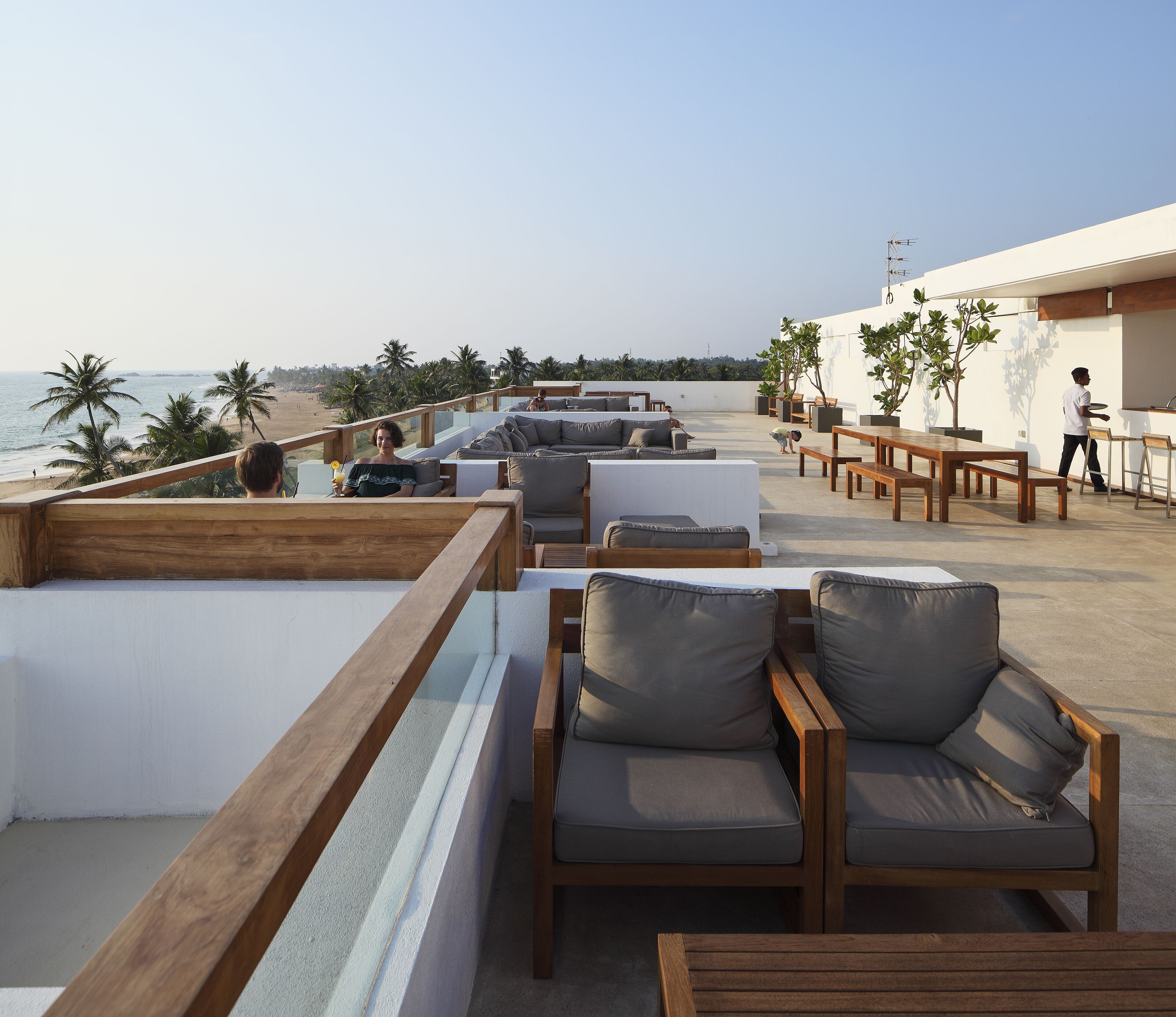 Eine Dachterrasse mit Sitzgelegenheiten im Freien, Blick auf den Strand und Palmen.