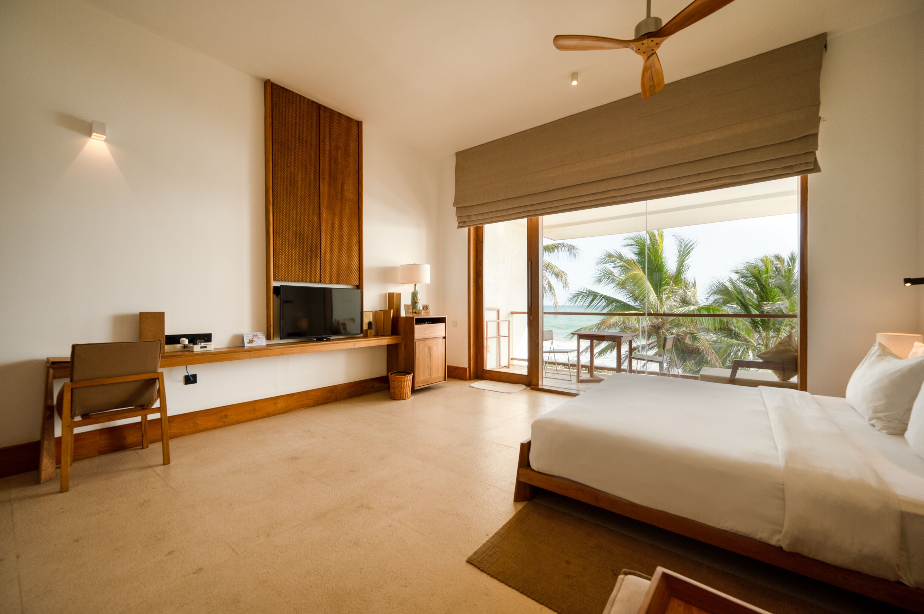 A hotel room with a bed, a desk with a TV, a chair, a window with a view of palm trees, and a ceiling fan.