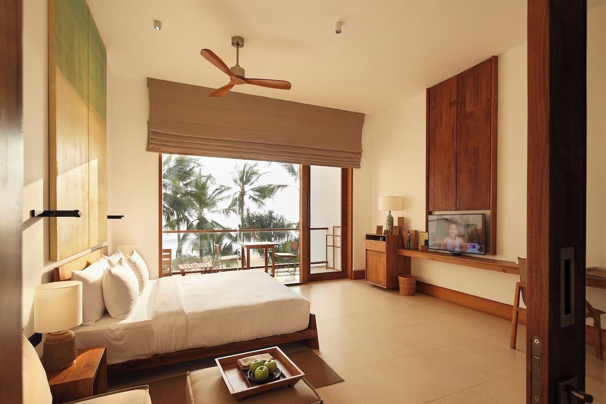 Quarto de hotel com cama, escrivaninha, televisão, ventoinha no teto e vista para palmeiras.