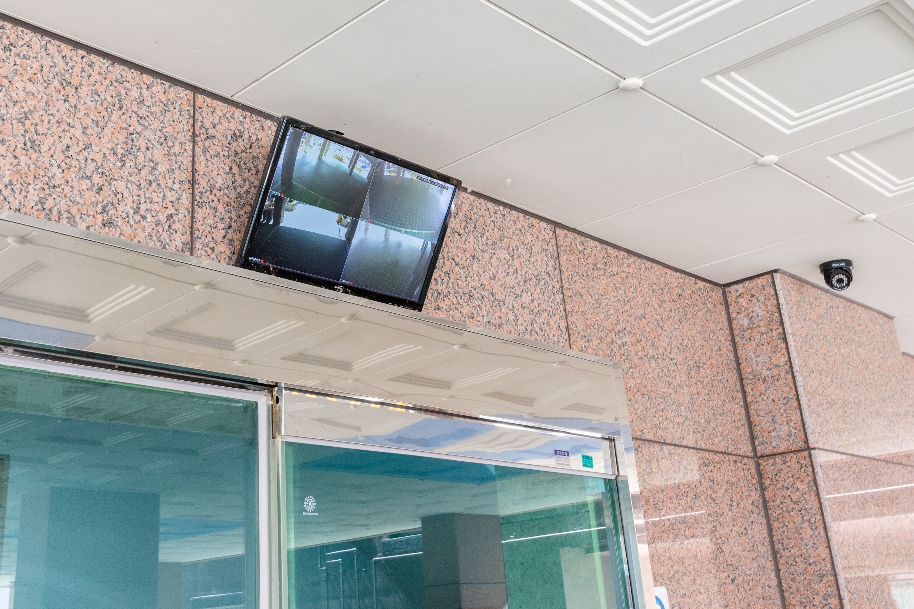 Une caméra de surveillance est fixée au plafond, au-dessus d’un mur recouvert de carreaux et d’une porte vitrée.