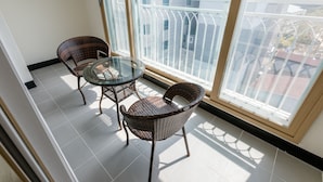 Un balcon avec deux fauteuils en osier et une table en verre, de grandes fenêtres offrant une vue sur les immeubles et un sol carrelé.