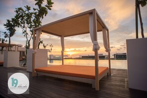 Una terraza de madera con una cama con dosel que da a una masa de agua y un atardecer.