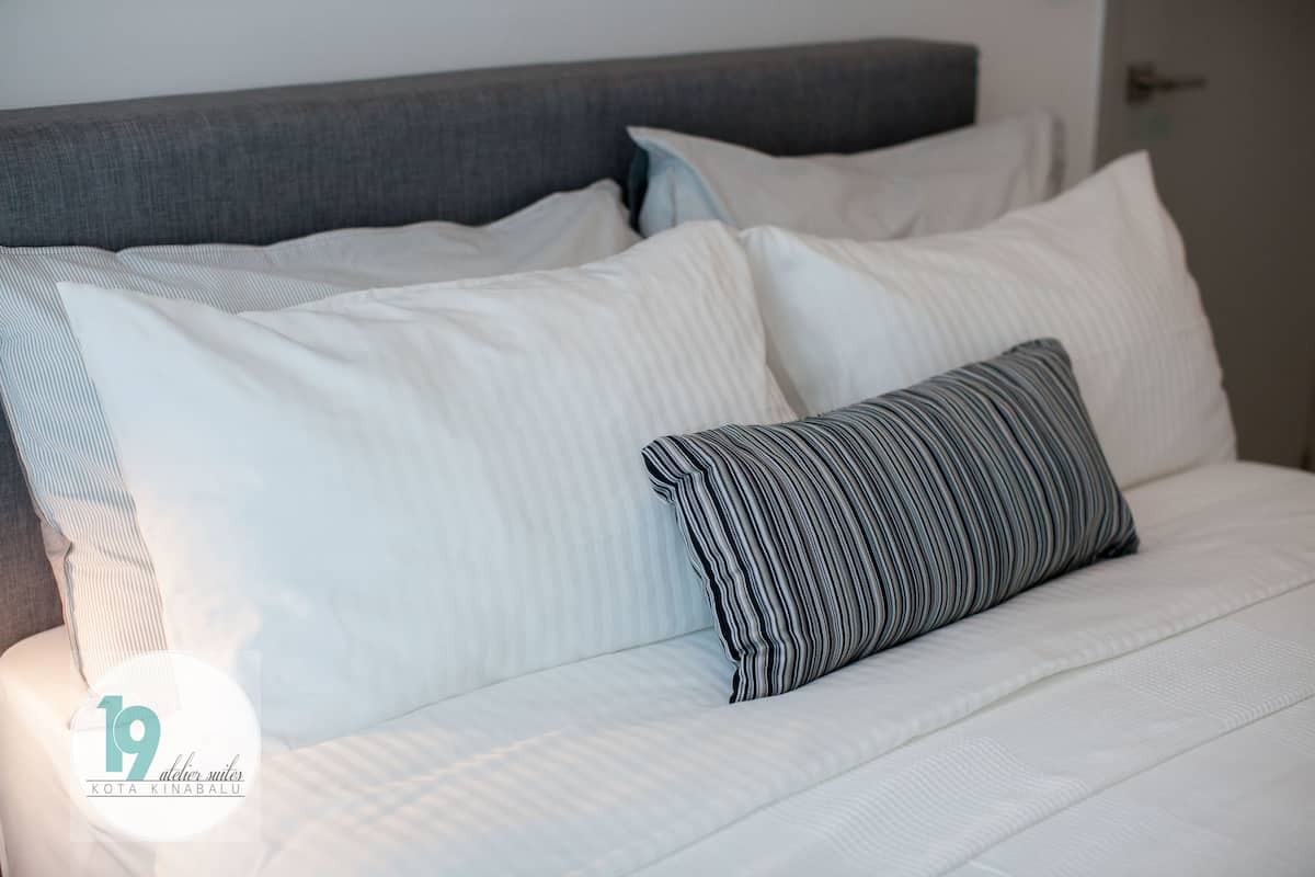 A neatly made bed with white bedding and striped pillows, a grey headboard, and a visible door in the background.
