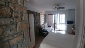 A room with a stone wall, a bed, a sofa, a TV, and a ceiling fan. - Kastro Konak Boutique Otel (Gökçeada)