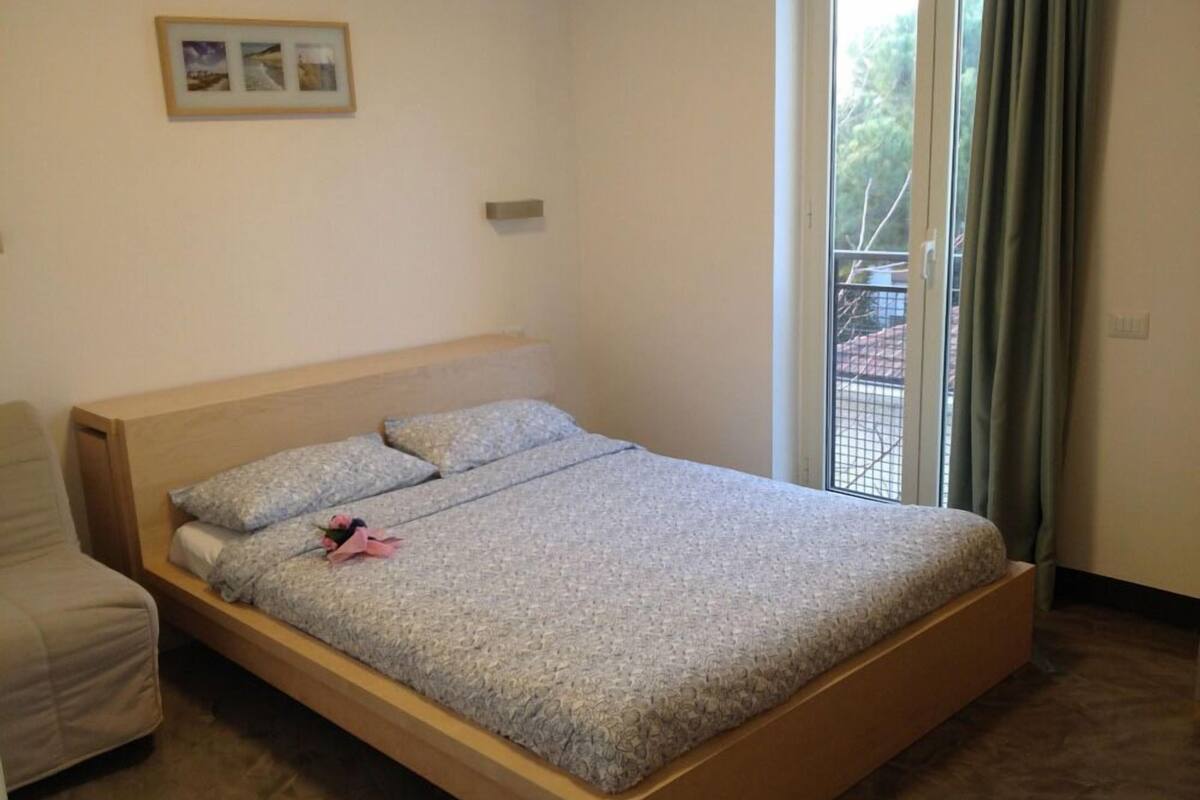 Ein Zimmer mit einem Einzelbett, einem Holzkopfende, einem kleinen Nachttisch und einem Fenster mit Vorhängen.