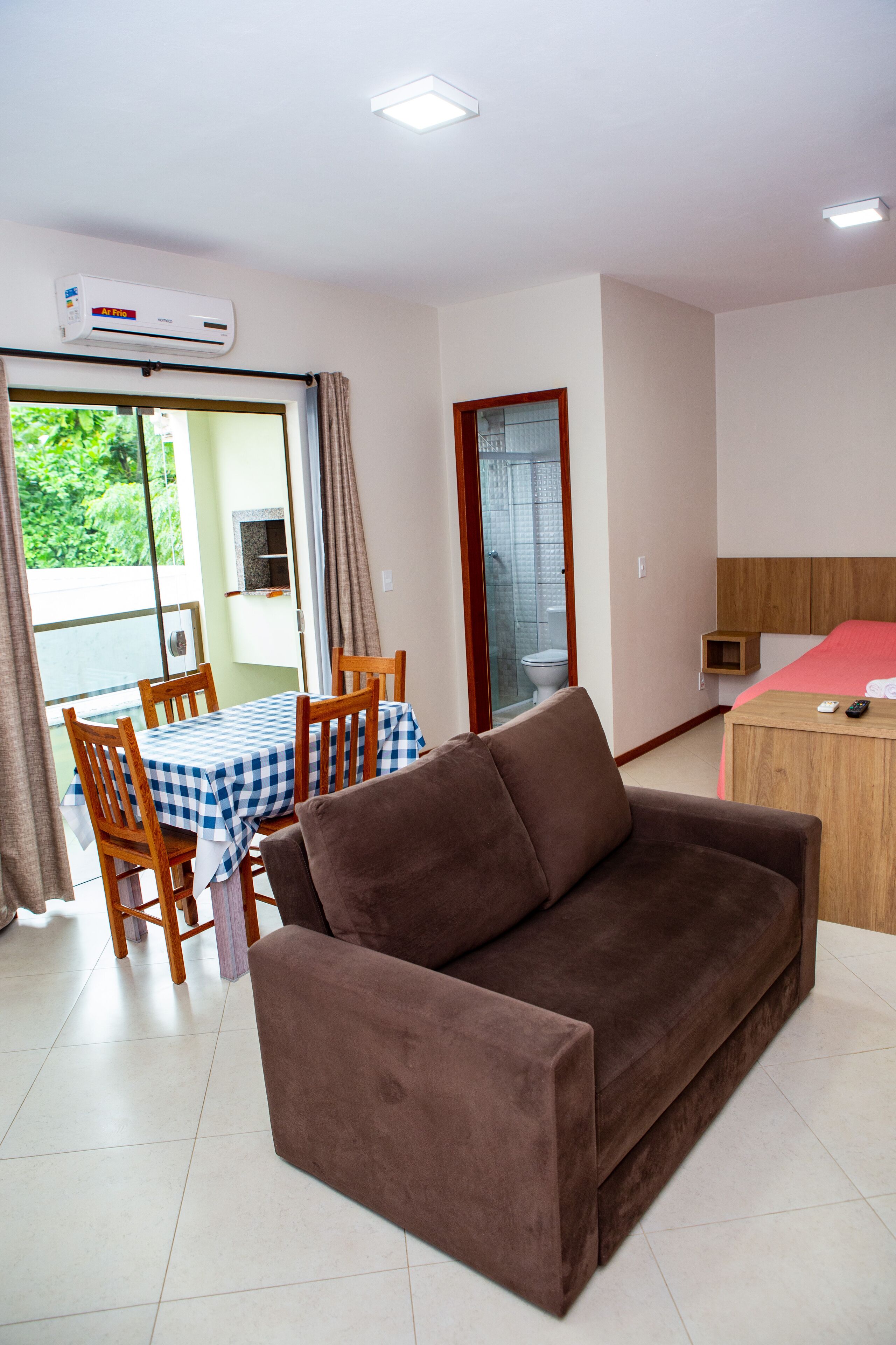 A furnished room with a brown sofa, a table with chairs, a bed, and a sliding glass door leading to an outdoor area.