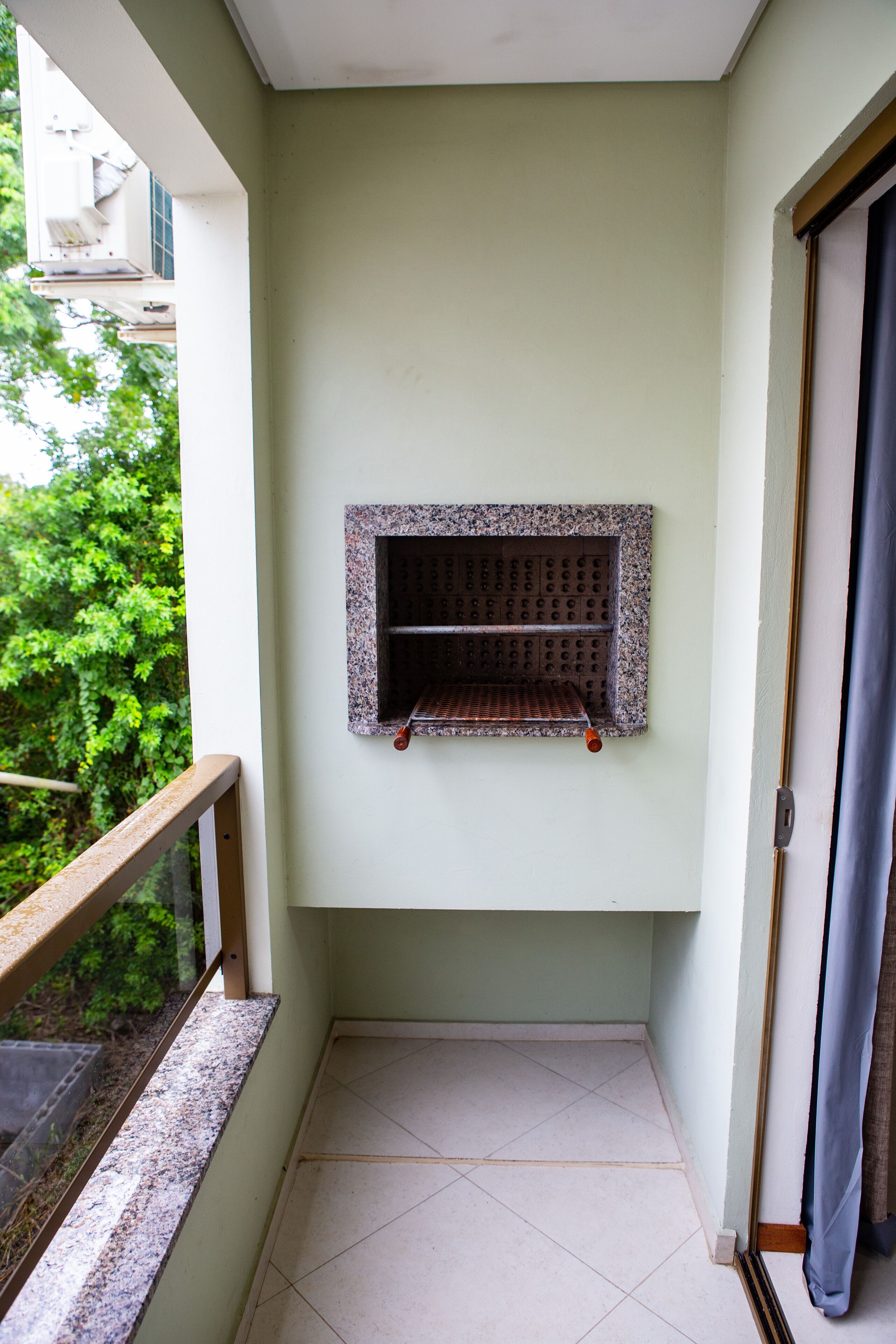 A small balcony with a tiled floor, a metal grill, and a view of greenery outside.