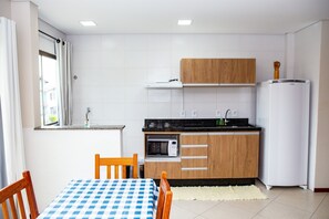 A compact kitchen with a microwave, stove, and refrigerator. There is a wooden cabinet above the stove and a window with curtains. A table with a checkered tablecloth and two chairs are in the foreground. - Canto Dos Passaros Flat Canasvieiras (Florianópolis)