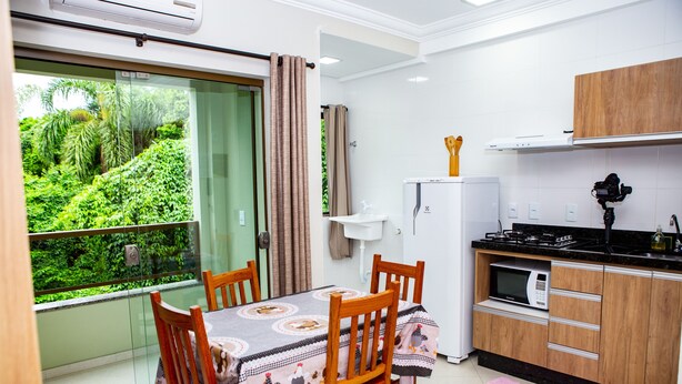 A modern kitchen with a dining area, a view of greenery, and a balcony. - Canto Dos Passaros Flat Canasvieiras (Florianópolis)