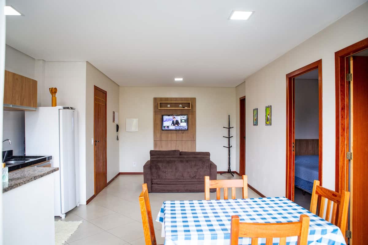 Een ruimtelijk ingerichte woonruimte met een kitchenette, een bank, een aan de muur bevestigde televisie en een eethoek met een tafel en stoelen.