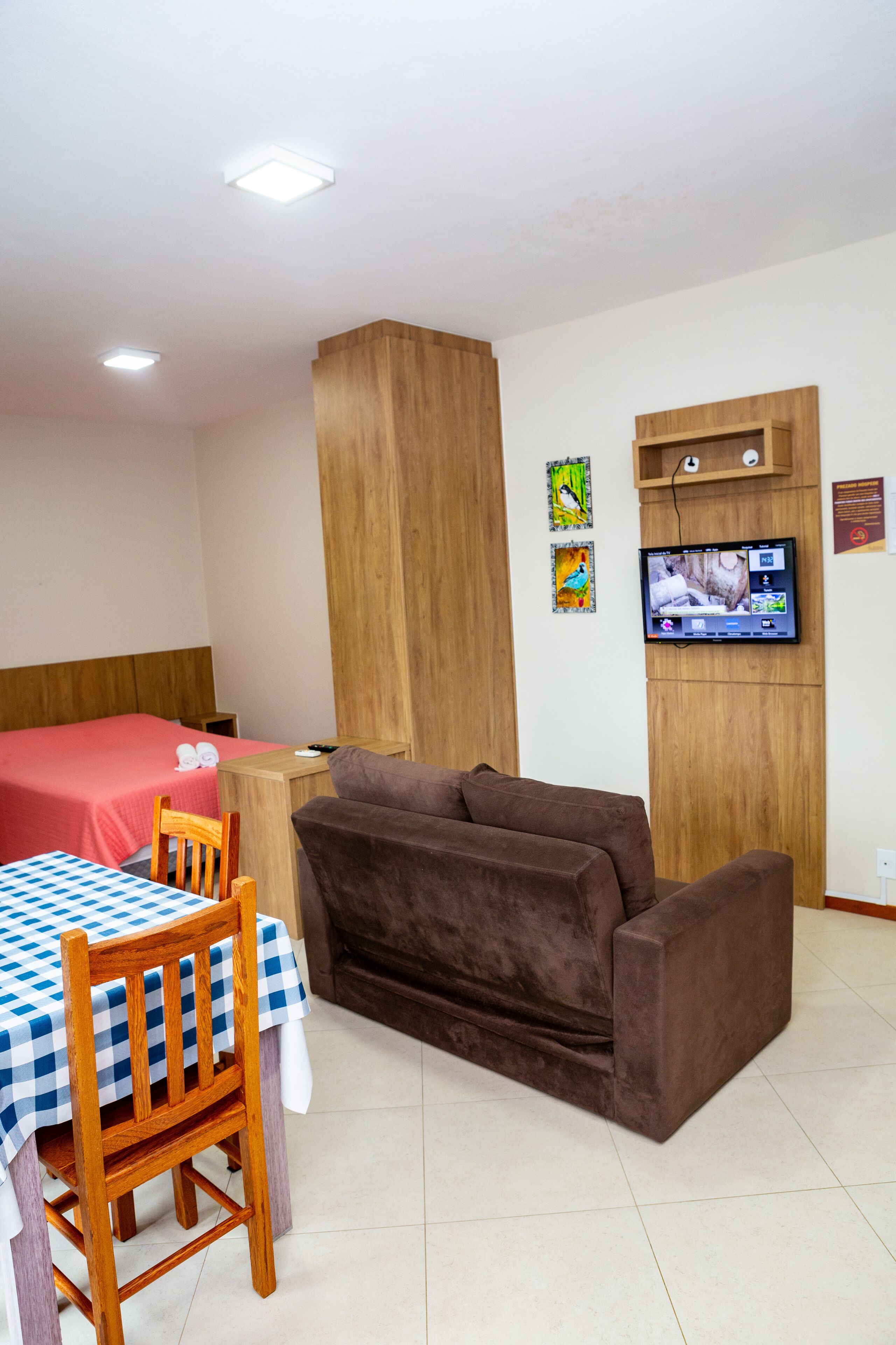 A small, furnished room with a bed, a brown sofa, a wooden chair, a table with a checkered tablecloth, a television mounted on the wall, and a small shelf with a power outlet.