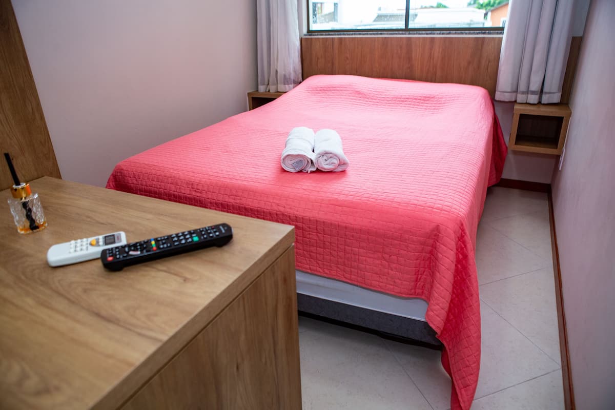 Een net opgemaakt bed met een roze dekbed en twee opgerolde handdoeken, een houten nachtkastje met een afstandsbediening, en een klein tafeltje met een fles.