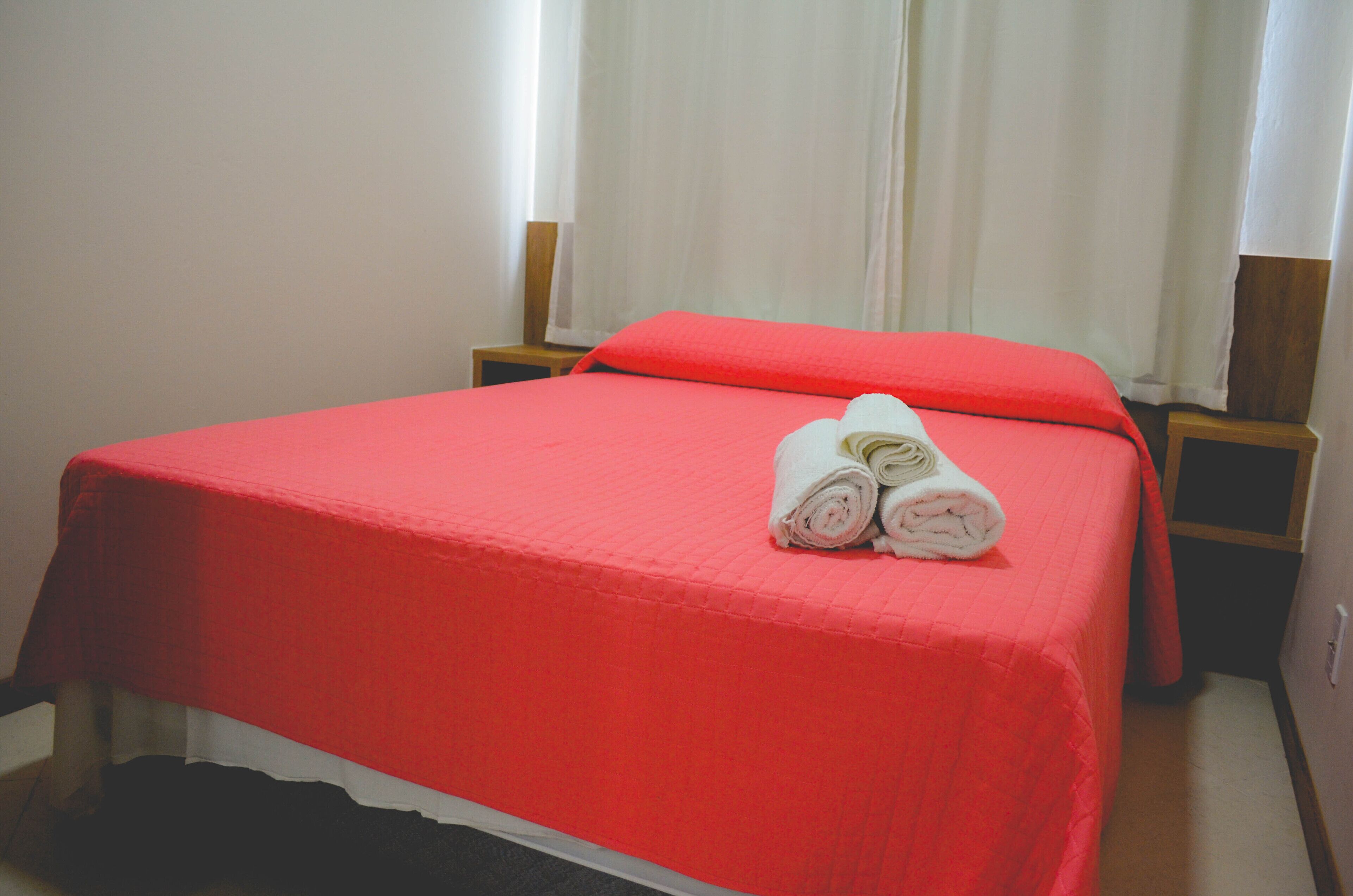 Un lit simple avec une couverture rouge, deux oreillers et des serviettes enroulées posées sur le lit.