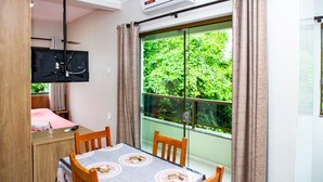 A room with a dining table, chairs, a TV mounted on the wall, and a large window with curtains. - Canto Dos Passaros Flat Canasvieiras (Florianópolis)