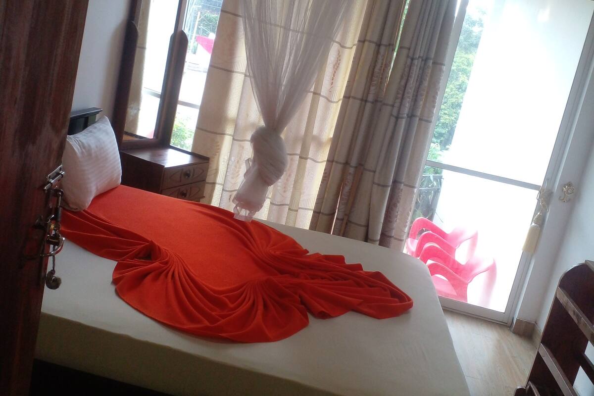 A bed with a red bedspread, a bedside table, a mirror, and a window with curtains.