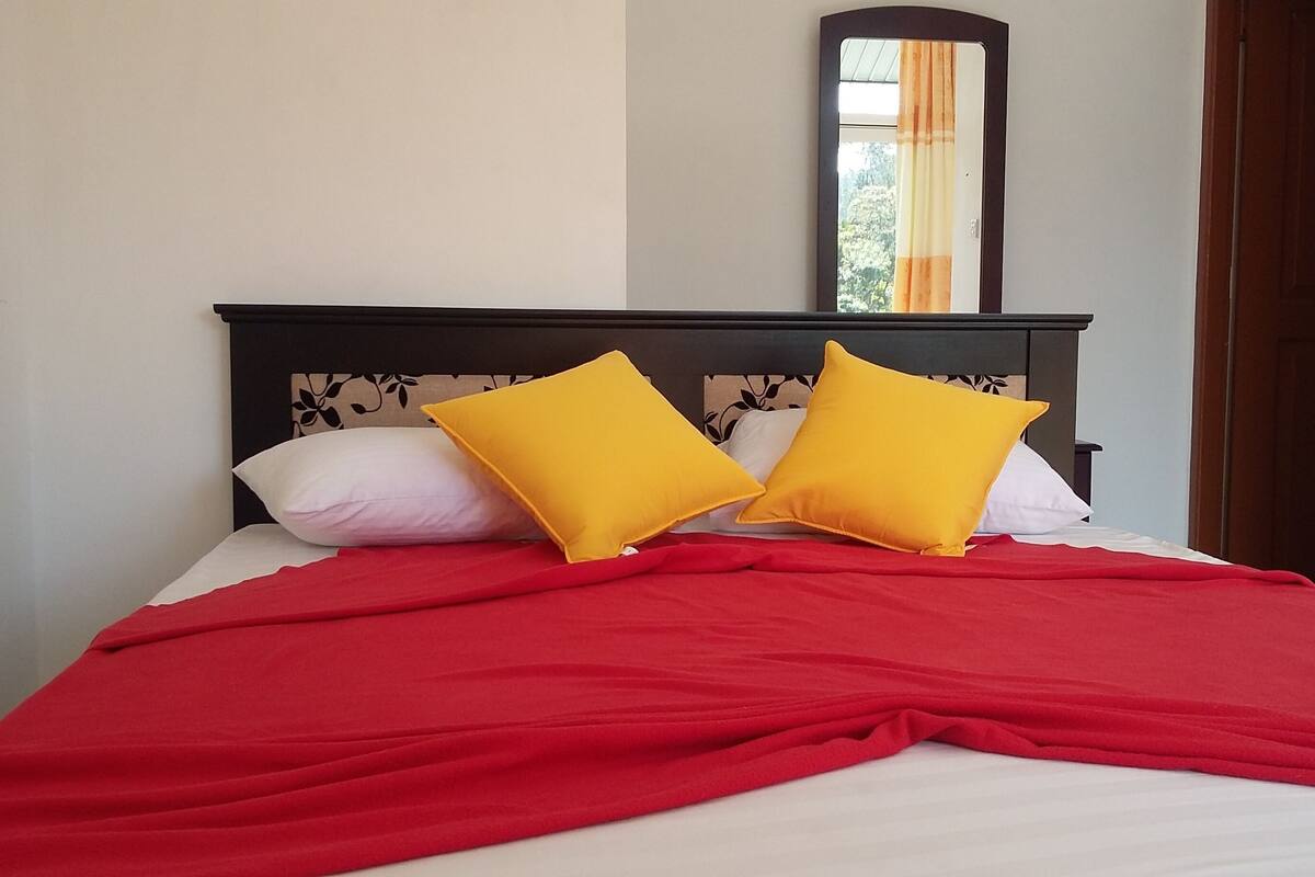 A bed with a red blanket and two yellow pillows, a dark wooden headboard, and a mirror on the wall.