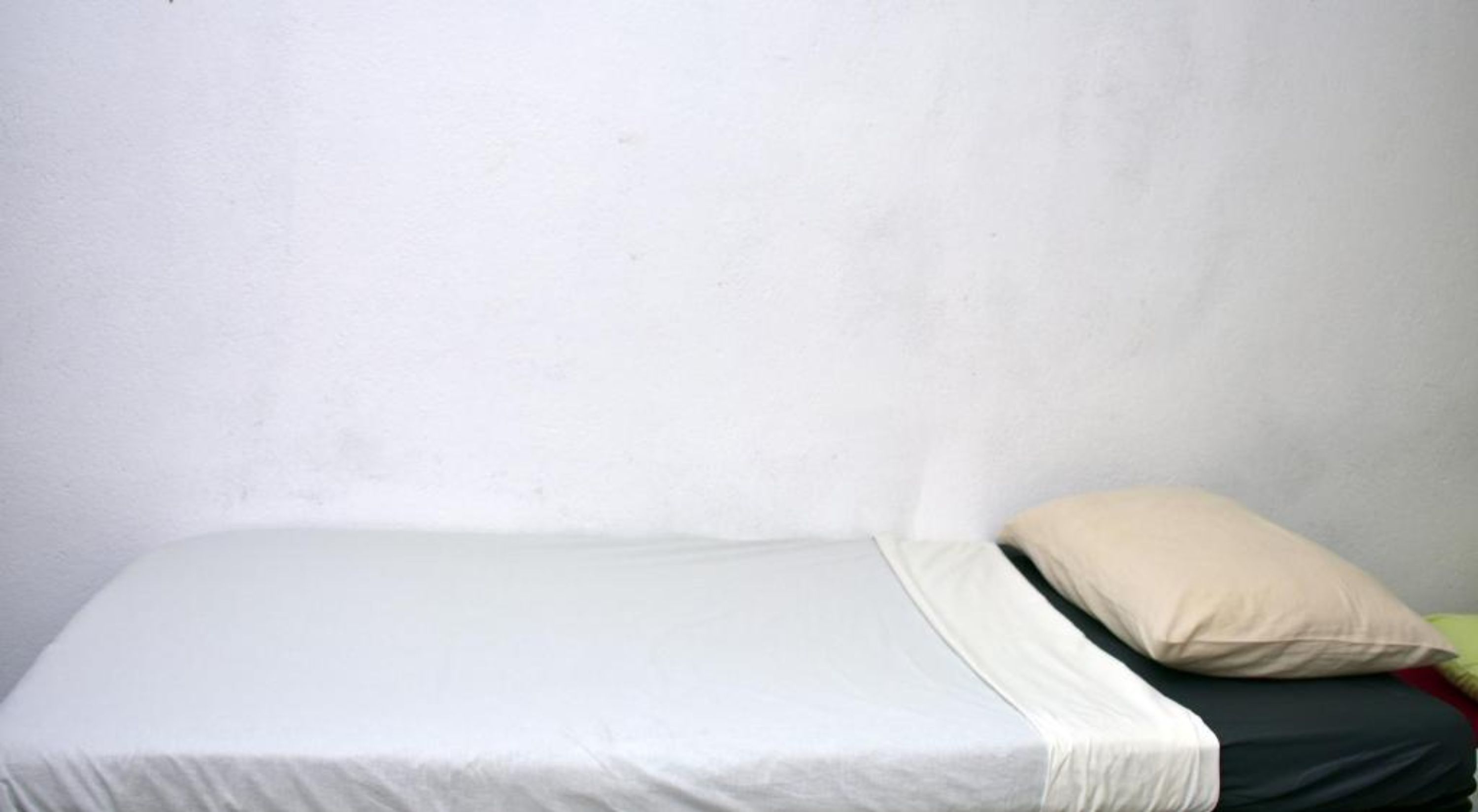 Een enkel bed met een witte matras en een beige kussen tegen een effen witte muur.