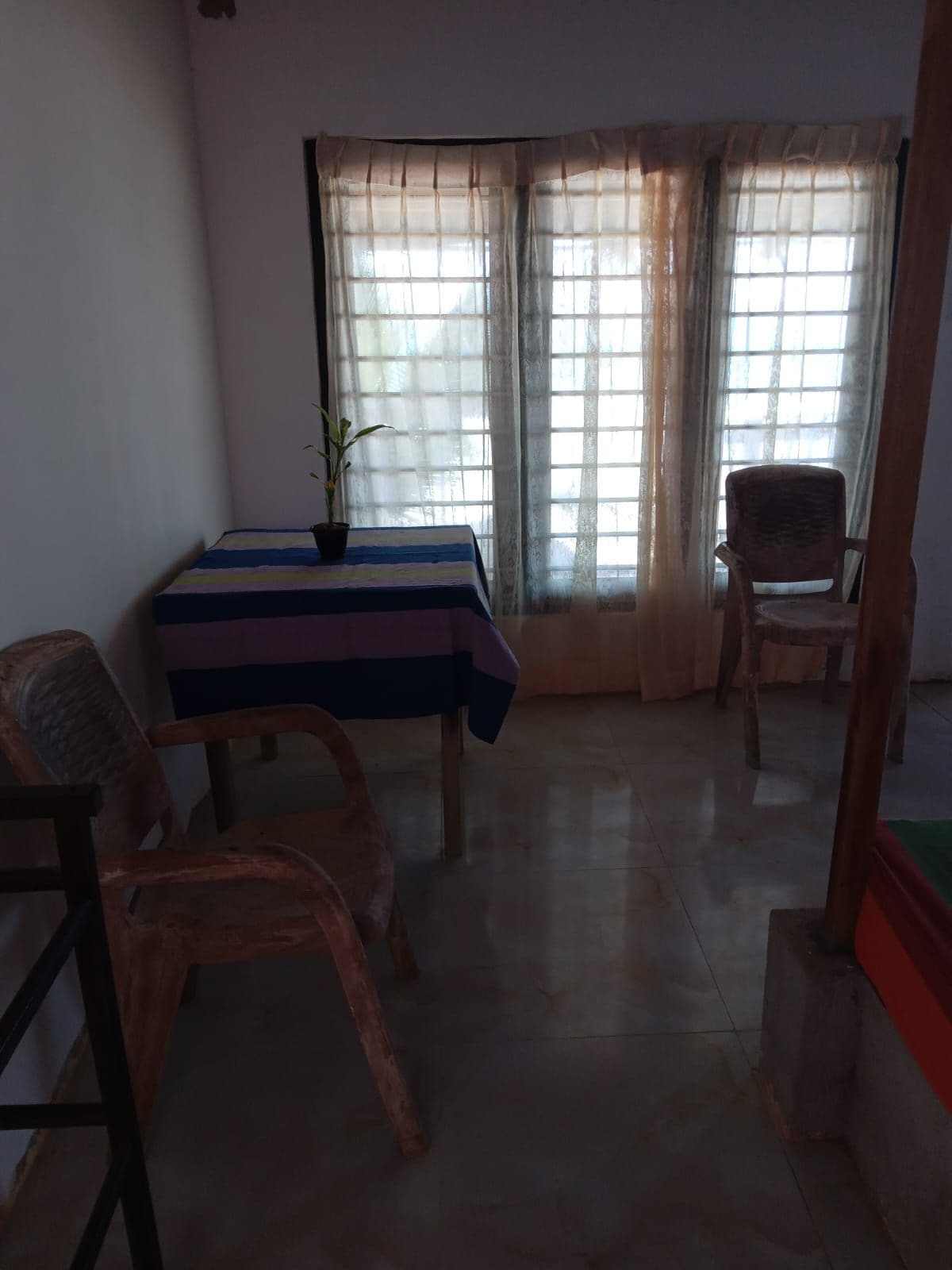 Una habitación con una mesa cubierta por un mantel a rayas, dos sillas de mimbre, una planta en maceta y una ventana con cortinas traslúcidas.