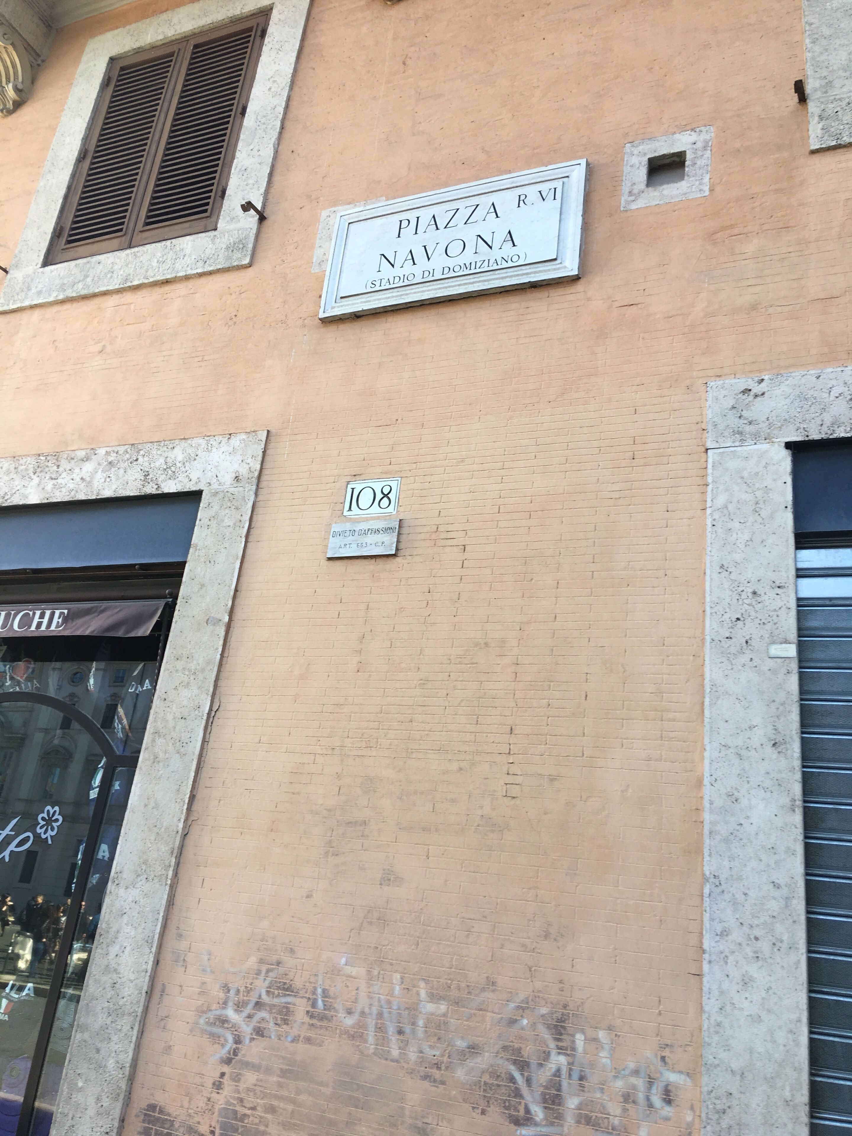 A building facade with a sign reading "PIAZZA R.VI NAVONA (STADIO DI DOMIZIANO)" and a smaller sign below stating "DIVIETO DAFFISSONI ART. 663-C.F.R."