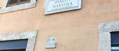 A building facade with a sign reading "PIAZZA R.VI NAVONA (STADIO DI DOMIZIANO)" and a smaller sign below stating "DIVIETO DAFFISSONI ART. 663-C.F.R."