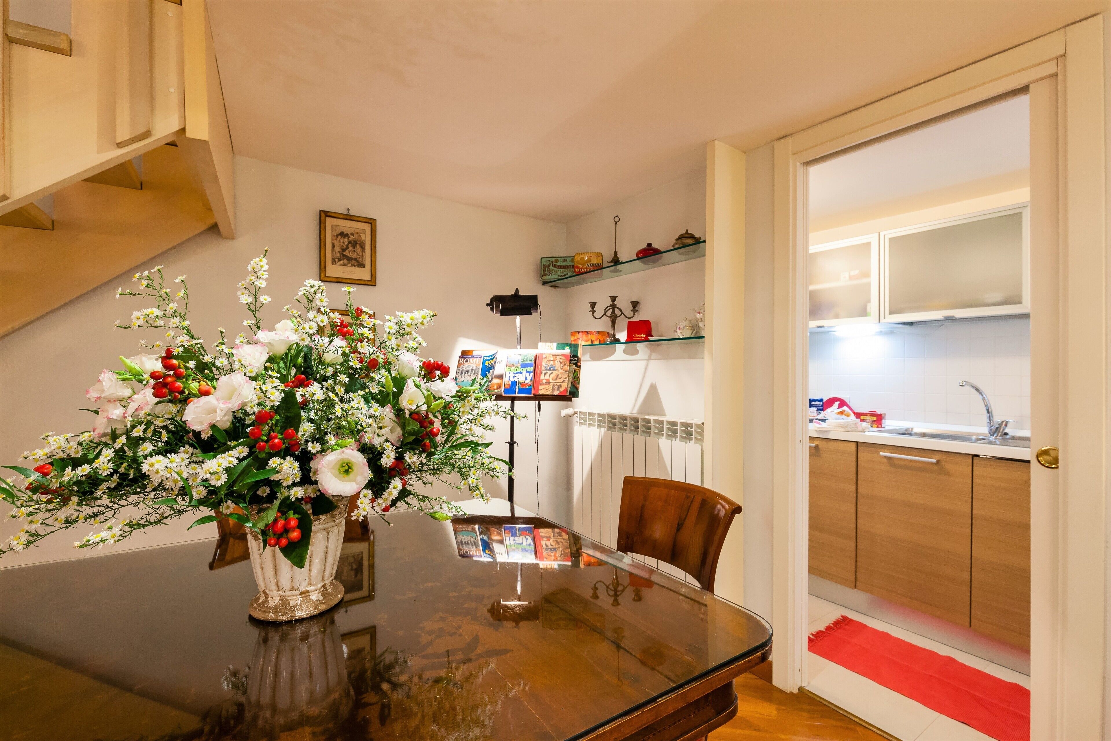 On aperçoit un coin repas avec une table en verre, un vase de fleurs et une chaise. Au fond, on distingue une cuisine avec des placards et un évier.