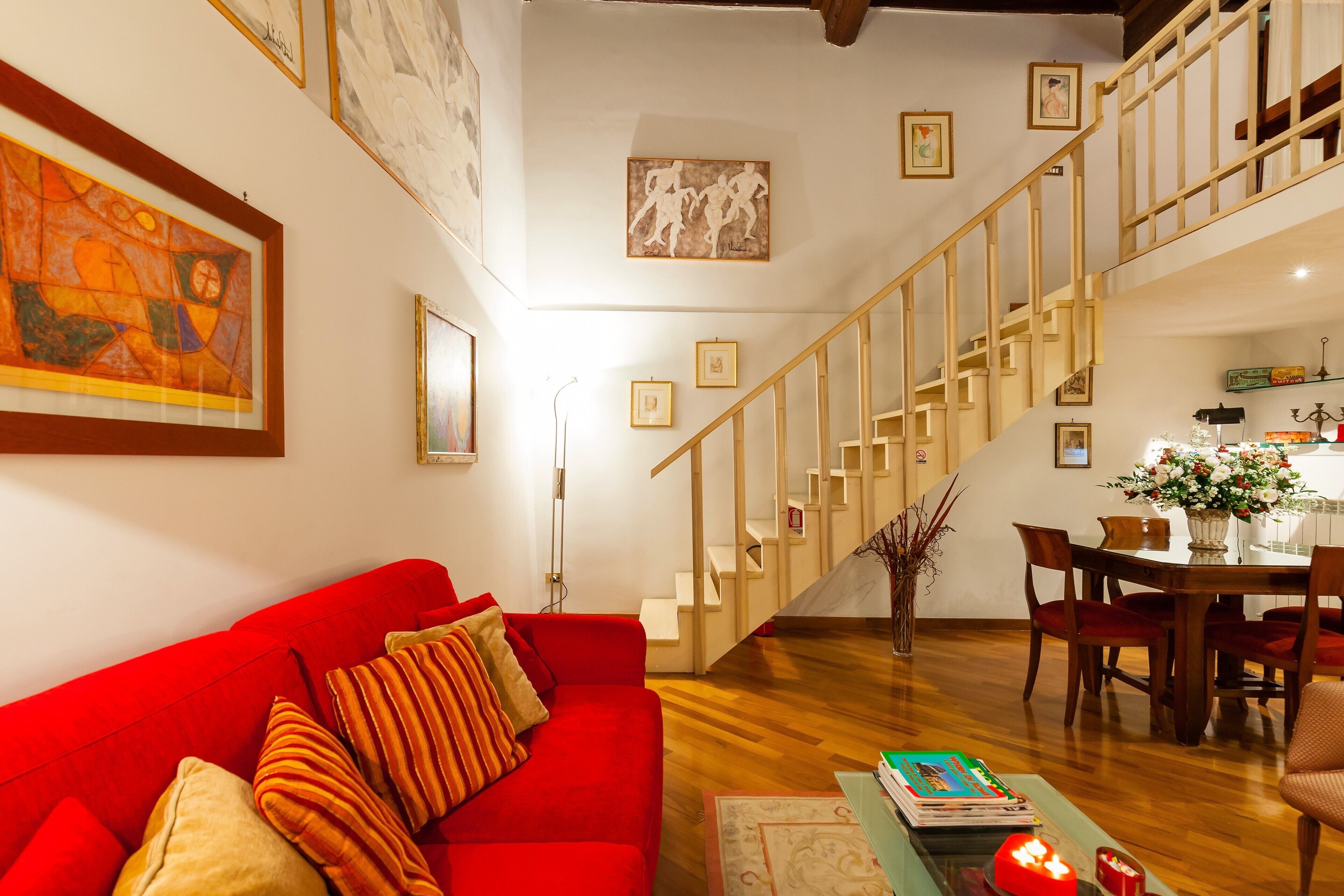 Un salon avec un canapé rouge, un escalier en bois, un coin repas et des œuvres d’art accrochées aux murs.