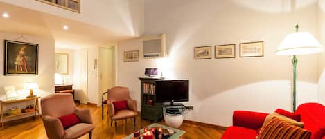 Uma sala de estar com um sofá vermelho, uma poltrona marrom, uma mesa de centro com livros e velas, uma televisão e quadros nas paredes.
